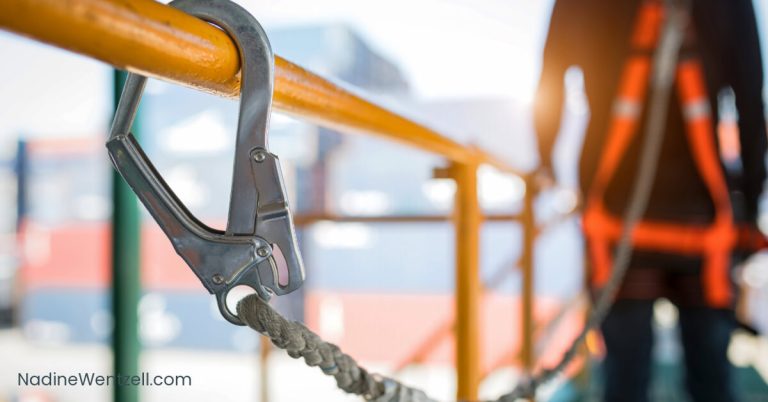 Close-up of a safety carabiner clipped to an orange railing, with a worker in a safety harness in the background. Emphasizes workplace safety.