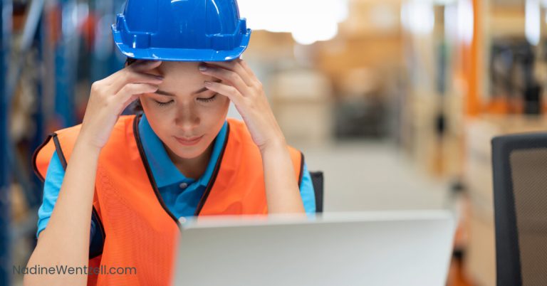 Female worker in a hard hat and safety vest sitting at a desk in a warehouse, holding her head with both hands in a stressed or focused posture.