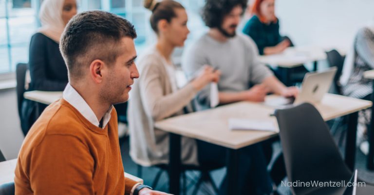 Group of adult learners seated at desks in a bright classroom, attentively engaging in a training session. Some are taking notes while others work on laptops.