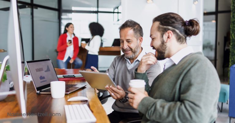 Two male coworkers collaborating at a desk with a tablet and coffee cup, while two female colleagues chat in the background in a modern office setting. Laptops and business charts are visible, indicating a professional workplace environment.