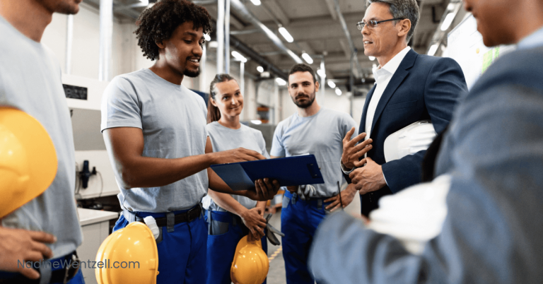 Group of industrial workers in uniform holding yellow hard hats, listening to a manager in a suit during a safety briefing on the factory floor. One worker holds a clipboard.
