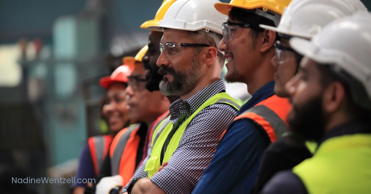 Diverse group of construction workers wearing safety vests and hard hats, listening attentively during a workplace safety meeting.