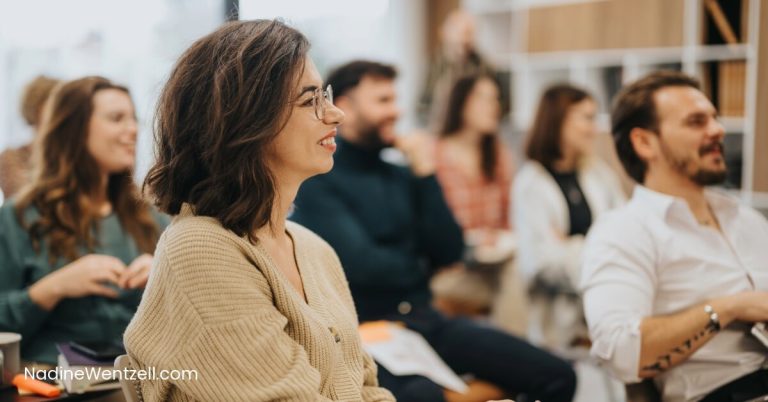 A group of adults attentively listens during a workshop or seminar. The focus is on a smiling woman with short brown hair and glasses wearing a beige sweater, seated among other participants in a bright, modern classroom setting. Everyone appears engaged and relaxed, suggesting a positive and interactive learning environment.